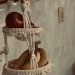 Handmade 3-tier macrame fruit hanging basket holding apples, bananas, and oranges in a kitchen corner by Kemet Vibes.